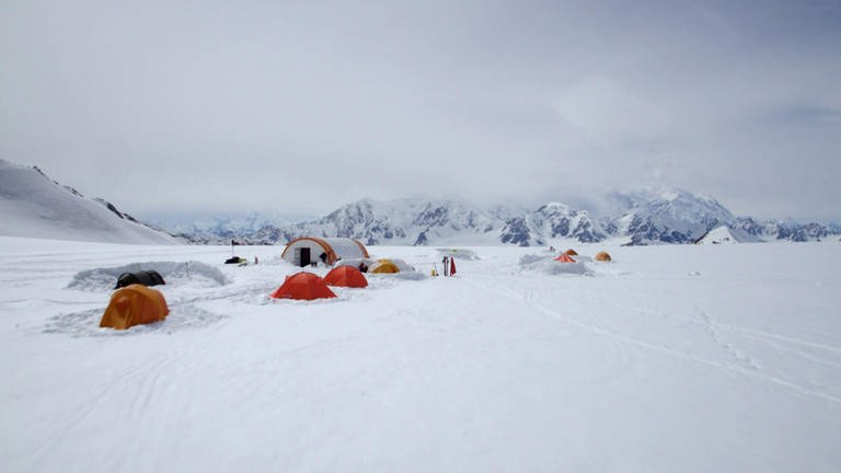 In einer schneebedeckten Ebene stehen einige große und kleine Zelte. Im Hintergrund sind steile, vernebelte Berge zu erkennen.