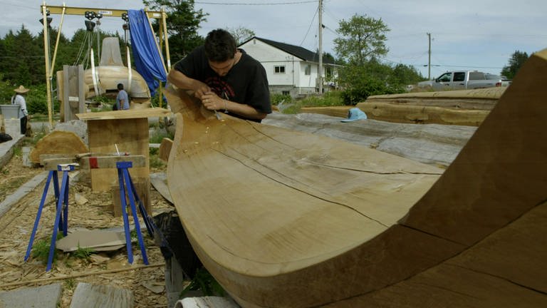 Ein Mann bearbeitet ein Holzboot, indem er Holzspäne von dem Stamm hobelt.