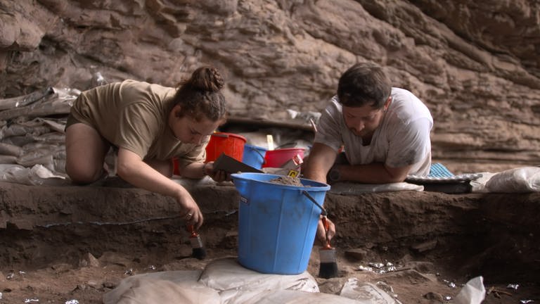Forscherin (links) und Forscher (rechts) knien an einer Ausgrabungsstätte und legen mit Pinseln vorsichtig Funde frei.