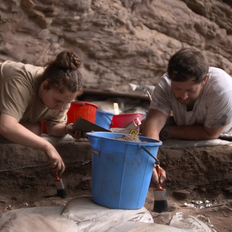 Forscherin (links) und Forscher (rechts) knien an einer Ausgrabungsstätte und legen mit Pinseln vorsichtig Funde frei.