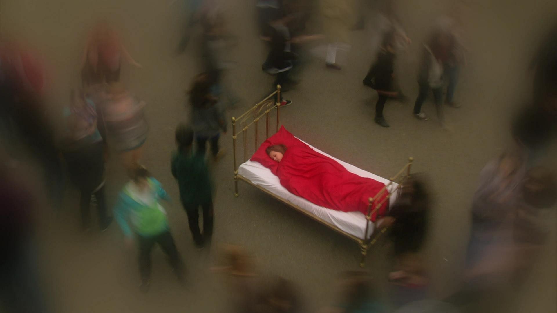 Eine Frau schläft in einem Bett auf einem Platz. Um sie herum laufen Menschen, die verschwommen abgebildet sind. (Foto: WDR)