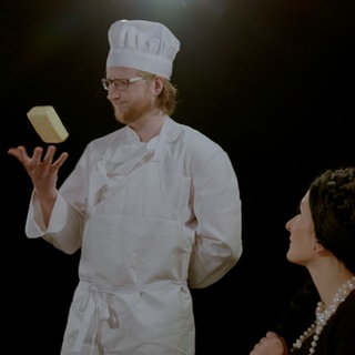 Eine sitzende Frau betrachtet einen Mann mit Kochmütze, der ein Stück Butter mit seiner rechten Hand hochwirft