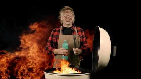 Ein Mann mit rußgeschwärztem Gesicht und Spiritusflasche in der Hand steht hinter einem Grill, aus dem eine Stichflamme schlägt.