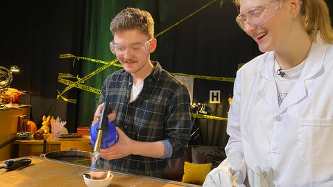 Ein Mann und eine Frau im Laborkittel mit Schutzbrillen richten die Flamme einer Lötlampe auf Plastikteile in einem Schmelztiegel