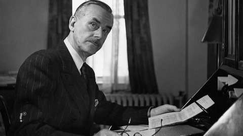 Thomas Mann sitzt an einem Schreibtisch und schaut in die Kamera