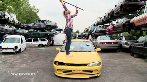 Die Moderatorin steht auf einem Auto und schlägt die Scheibe ein.