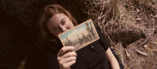 Spielszene: Joan Hinton hält lächelnd eine Postkarte in der Hand. Die Kernphysikerin arbeitet später am Manhattan-Projekt – der Entwicklung der Atombombe.