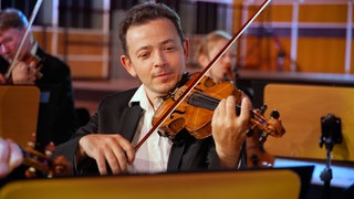 Ein Mann spielt Violine, im Hintergrund weitere Mitglieder der Streichergruppe.