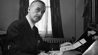 Thomas Mann sitzt an einem Schreibtisch und schaut in die Kamera