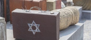 Ein Mahnmal für die Deportationen von Juden in Konzentrationslager während des Nationalsozialismus: Alte Koffer stehen auf der Straße. Auf einem Koffer ist ein Davidstern, der Griff ist aus Stacheldraht.