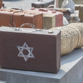 Ein Mahnmal für die Deportationen von Juden in Konzentrationslager während des Nationalsozialismus: Alte Koffer stehen auf der Straße. Auf einem Koffer ist ein Davidstern, der Griff ist aus Stacheldraht.