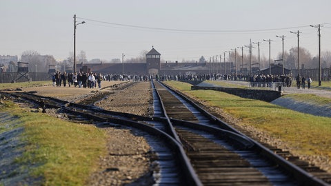 Frontalansicht des Konzentrationslagers Auschwitz von den Bahngleisen. Die Gleise führen zum Eingang. Auf dem Gelände laufen viele Besucher. Die ankommenden Häftlinge wurden noch vor dem Lager in zwei Gruppen eingeteilt – die Selektion.
