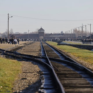 Frontalansicht des Konzentrationslagers Auschwitz von den Bahngleisen. Die Gleise führen zum Eingang. Auf dem Gelände laufen viele Besucher. Die ankommenden Häftlinge wurden noch vor dem Lager in zwei Gruppen eingeteilt – die Selektion.