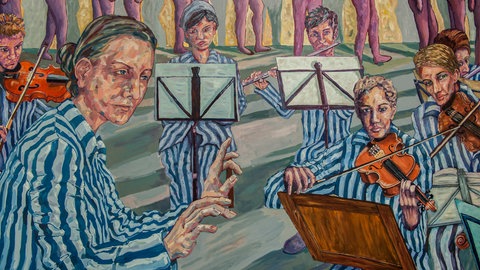 Ein Gemälde zeigt KZ-Häftlinge in blau-weiß gestreifter Kleidung. Eine der Frauen dirigiert, die anderen sitzen teils hinter Notenständern und spielen Geige. Das Gemälde stellt das Mädchenorchester in Auschwitz dar.