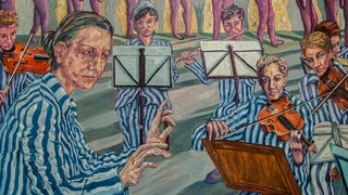 Ein Gemälde zeigt KZ-Häftlinge in blau-weiß gestreifter Kleidung. Eine der Frauen dirigiert, die anderen sitzen teils hinter Notenständern und spielen Geige. Das Gemälde stellt das Mädchenorchester in Auschwitz dar.