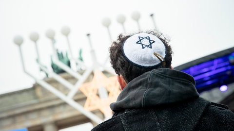 Ein Mann mit schwarzer Jacke von hinten. Auf dem Kopf trägt er eine weiße Kippa mit einem blauen Davidsstern – ein Symbol des Judentums. Im Hintergrund ein Chanukka-Leuchter. Es gibt wieder mehr Fälle von Antisemitismus in Deutschland.