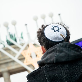 Ein Mann mit schwarzer Jacke von hinten. Auf dem Kopf trägt er eine weiße Kippa mit einem blauen Davidsstern – ein Symbol des Judentums. Im Hintergrund ein Chanukka-Leuchter. Es gibt wieder mehr Fälle von Antisemitismus in Deutschland.