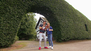 Ein Mann und eine Frau stehen vor einer sehr hohen Hecke und schauen auf einen Kompass.