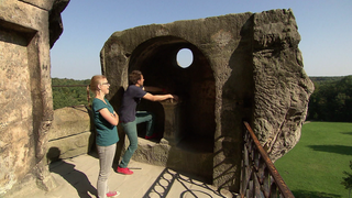 Ein Mann und eine Frau stehen auf einer Steinmauer und gucken durch ein Felsloch über grüne Felder.