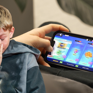 Im Vordergrund ein Junge, der frustriert nach unten schaut; im Hintergrund ein Handyspiel, bei dem gerade etwas gekauft wird