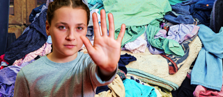 Im Vordergrund ein Mädchen in Abwehrhaltung, im Hintergrund ein großer Haufen bunter Wäsche