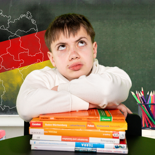 Junge mit fragendem Blick sitzt vor einer Schultafel mit Deutschlandumriss, nach Bundesländern aufgeteilt, darüber die Farben Schwarz-Rot-Gold.