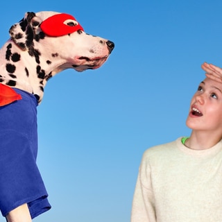 Links ein Hund im Superheldenkostüm mit großem rotem Umhang, rechts ein erstauntes Mädchen