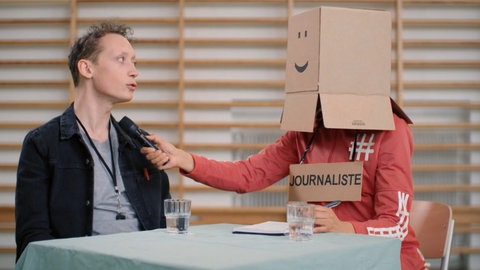 Moderator Jonas erzählt einer Person mit einem Karton über dem Kopf und einem Mikro eine Geschichte. Wie unterscheidet man das Imperfekt von anderen französischen Vergangenheitsformen?