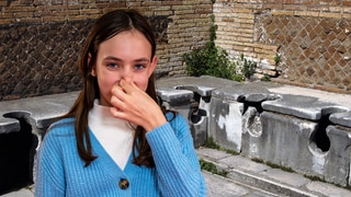 Im Hintergrund die Ruinen einer Toilettenanlage der alten Römer; im Vordergrund ein Mädchen, das sich die Nase zuhält