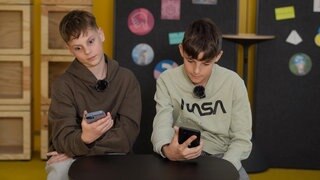 Zwei Schüler sitzen in einem modernen Klassenzimmer. Beide schauen genervt auf ihr Smartphone.