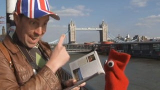 Ein Mann trägt eine Kappe, die mit dem Unionjack bedruckt ist. Neben ihm eine rote Strumpfhandpuppe. Sie stehen vor der Tower Bridge in London.