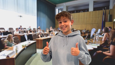 Junge zeigt Daumen hoch, im Hintergrund ein Sitzungssaal mit Abgeordneten. In Jugendparlamenten können sich Schüler in die Politik einbringen.