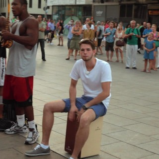 Innenstadt: Ein junger Mann sitzt auf einem Cajón. Um ihn herum sind viele Menschen.