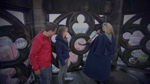 Drei Personen stehen auf dem Turm der Lambertikirche in Münster. Eine Frau spielt Horn.