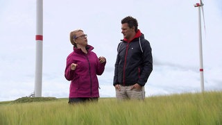 Ein Mann und eine Frau stehen im Dünengras vor Windrädern.
