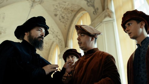 Vier Männer in alten Gewändern unterhalten sich in einem prächtigen Raum mit hohen Decken und Stuck