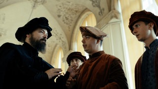 Vier Männer in alten Gewändern unterhalten sich in einem prächtigen Raum mit hohen Decken und Stuck