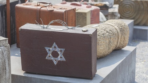 Ein Mahnmal für die Deportationen von Juden in Konzentrationslager während des Nationalsozialismus: Alte Koffer stehen auf der Straße. Auf einem Koffer ist ein Davidstern, der Griff ist aus Stacheldraht.