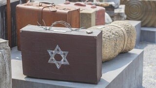 Ein Mahnmal für die Deportationen von Juden in Konzentrationslager während des Nationalsozialismus: Alte Koffer stehen auf der Straße. Auf einem Koffer ist ein Davidstern, der Griff ist aus Stacheldraht.