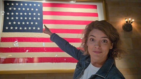 Host Lena Ganschow zeigt auf die amerikanische Flagge.