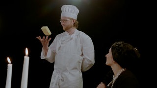 Eine sitzende Frau betrachtet einen Mann mit Kochmütze, der ein Stück Butter mit seiner rechten Hand hochwirft