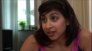 Eine junge Frau sitzt in einer Küche und spricht mit hochgezogenen Augenbrauen.