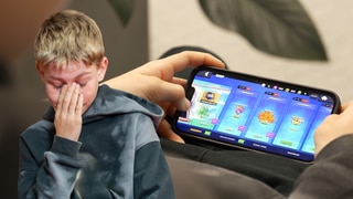 Im Vordergrund ein Junge, der frustriert nach unten schaut; im Hintergrund ein Handyspiel, bei dem gerade etwas gekauft wird