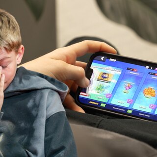 Im Vordergrund ein Junge, der frustriert nach unten schaut; im Hintergrund ein Handyspiel, bei dem gerade etwas gekauft wird