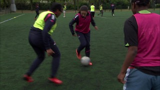 Drei Menschen in roten und gelben Leibchen stehen auf einem Fußballplatz und spielen miteinander Fußball.