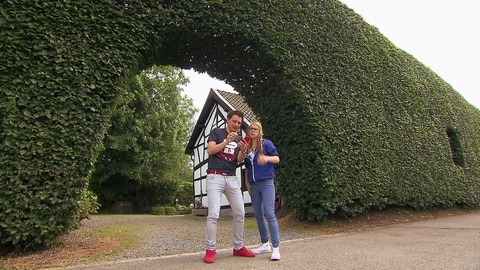 Ein Mann und eine Frau stehen vor einer sehr hohen Hecke und schauen auf einen Kompass.