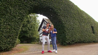 Ein Mann und eine Frau stehen vor einer sehr hohen Hecke und schauen auf einen Kompass.