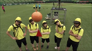 Gelb gekleidete Personen stehen auf einem Sportplatz und beobachten ein Pendel.