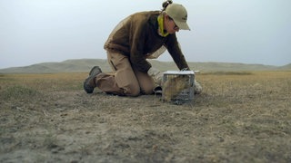 Eine Frau kniet im Staub, neben ihr steht ein kleiner Käfig, in dem ein Präriehund gefangen wurde.