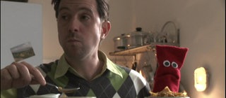 Ein Mann und eine rote Strumpfhandpuppe sitzen am Tisch und frühstücken.